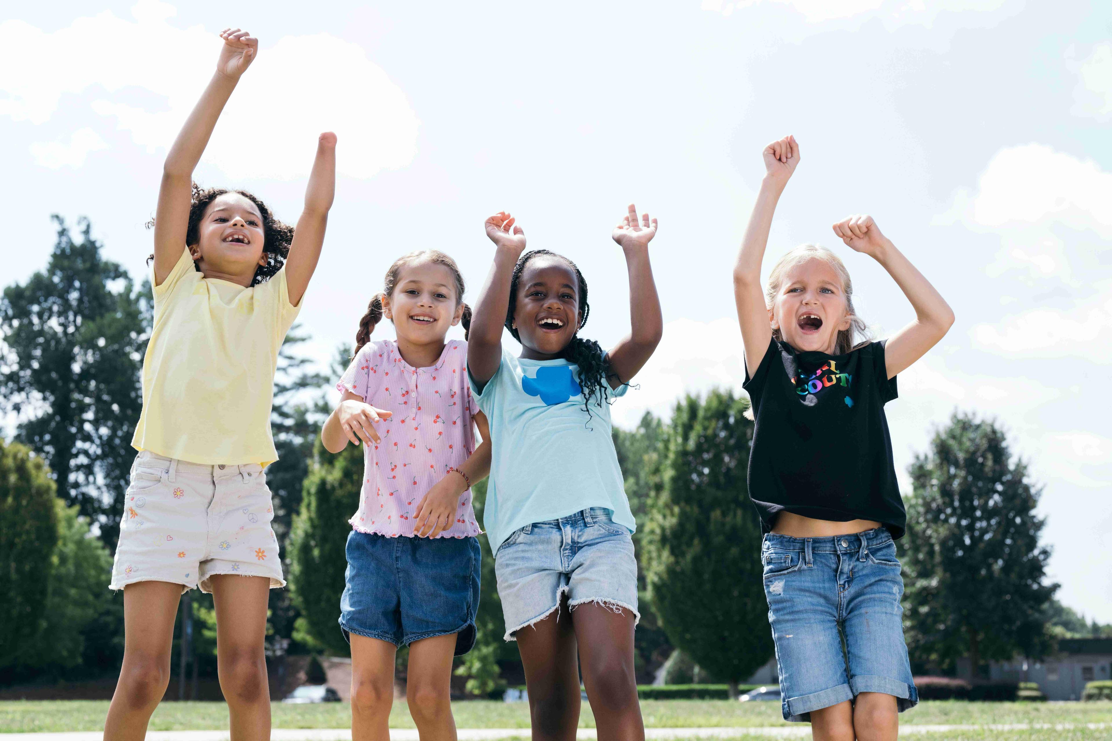 A group of Girl Scouts spending time outdoors and jumping in the air with their arms raised above their heads.